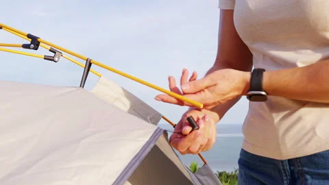 Bare Hands Attach a Yellow Pole to a Beige Dome Tent Beside Calm Water and Green Stock Footage 348753282