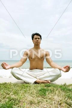 Barechested Man Sitting In Lotus Position, Ocean In Background, Full Length