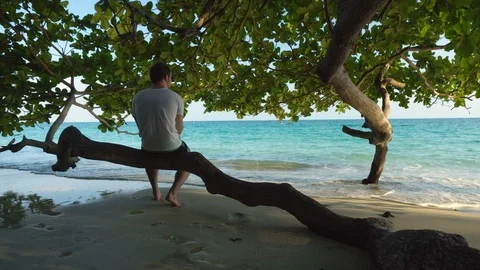 Barefoot Man Sitting Below Tree at Tropical Beach Walks to Ocean To Rinse Hands Stock Footage 119692713