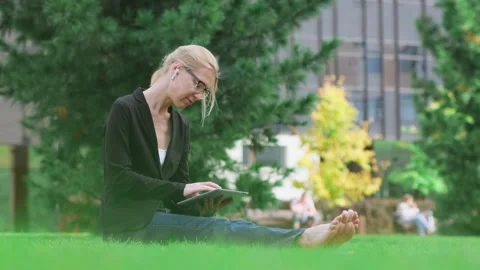 Barefoot woman using a tablet sitting on green lawn in city park. Stock Footage 221768078
