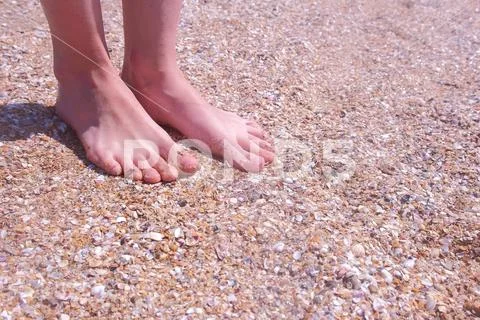 Barefooted girl traveller massaging feet on sandy beach in seashells at seaside. Stock Photos