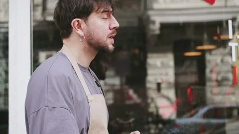 Barista on break outside cafe with cigarette while colleagues serve customer in Stock Footage 330978362