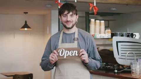 Barista welcomes customer to cafe and shows the open sign with colleagues Stock Footage 330977940