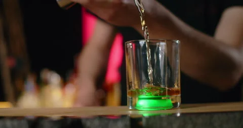 Barman pours portion of alcohol from jigger to the glass with green backlight in Stock Footage 156104061