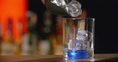 Bartender adds ice cubes to the glass with blue backlight in slow motion, making Stock Footage 156263950