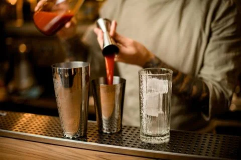 Bartender masterfully pours red drink from metal jigger into shaker on bar 스톡 사진