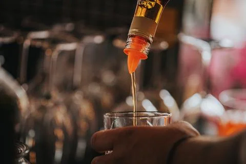 Bartender pouring amber liquid from a bottle into a glass, with blurred bar.. Stock Photos