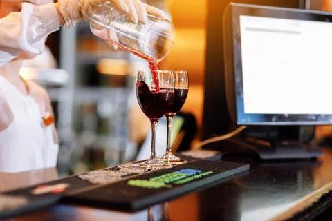 Bartender is pouring red wine into two long-stemmed wineglasses from a measur Stock Photos