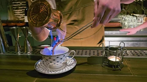 Bartender pours flaming liquid from a copper pot into a patterned teacup, with Stock Footage 340264448