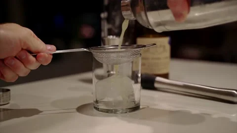Basil gimlet strained in slow motion over a large ice cube Stock Footage 221592045