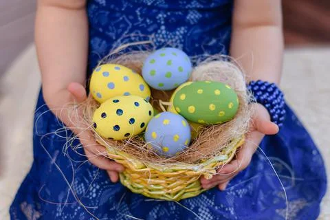 A basket with colorful Easter eggs in the hands of a little girl in a blue dress Stock Photos