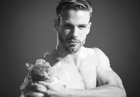 Bath time. Serious bearded man washing body with soapy sponge and moisturizin Stock Photos