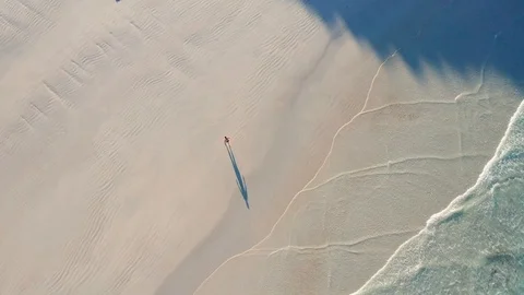 Bather walks up white beach casting shadow in early morning light after swim Stock Footage 125453318