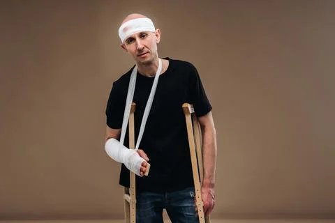A battered man with a bandaged head and a cast on his arm stands on crutches on Stock Photos