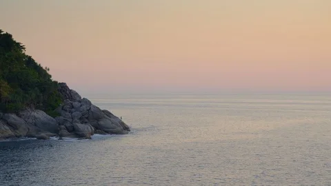 Beach with big sharp stones, hill and forest in warm sunset and fisherman Stock Footage 114917702