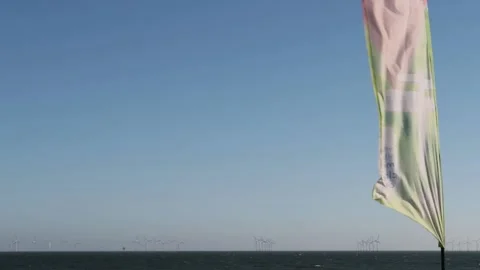 Beach flag flapping  in wind. Offshore wind farm turbines in background Stock Footage 173482810