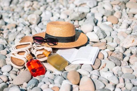 Beach hat on opened book with sunscreen and shoes on pebble beach Stock Photos
