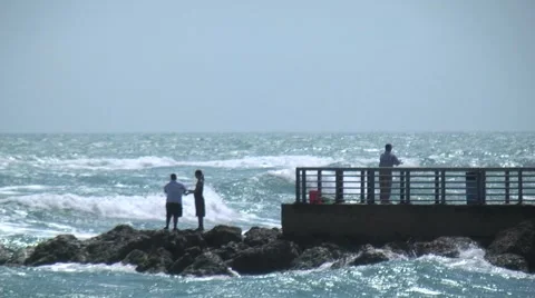 Beach scenic people fishing at end of pier on the rocks with waves crashing a Stock Footage