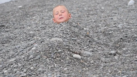 Beach summer vacation. Boy gets out from under the pebbles smiles and shows the Stock Footage 119598214
