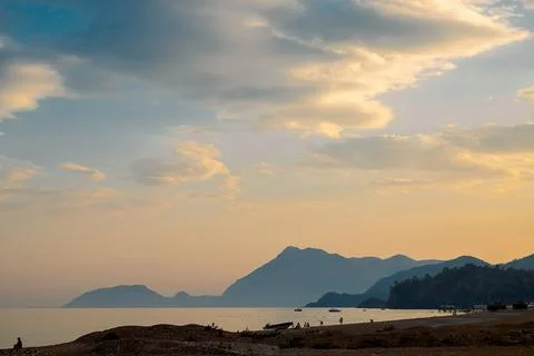 Beach at sunset while boats float on the water. Mountains rise in the backgro Stock Photos