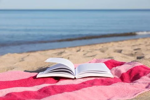Beach towel and open book on sand near sea, space for text Stock Photos