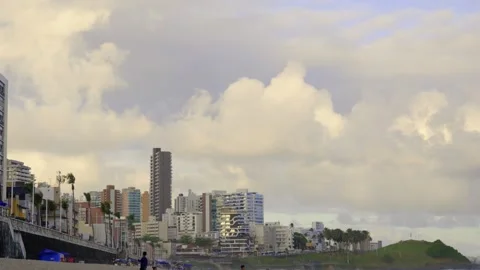 Beachgoers walking on Farol da Barra beach with coastal city skyline under cloud Stockbeeldmateriaal 319207342