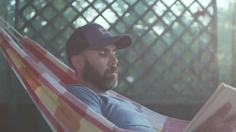 Beard caucasian man is reading a book in a hammock on the terrace in a sunset li 스톡 동영상 200917219