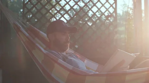 Beard caucasian man is reading a book in a hammock on the terrace in a sunset li Stock Footage 200917430