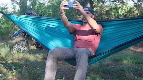 Beard man in a cap is sitting in a hammock in the forest near a motorcycle and d Stock Footage 214312364