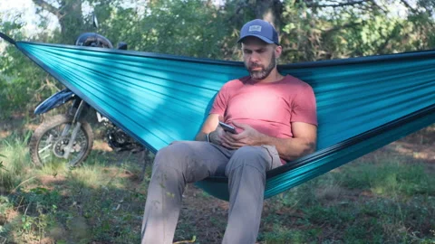 Beard man in a cap is sitting in a hammock in the forest near a motorcycle and w Stock Footage 214313654