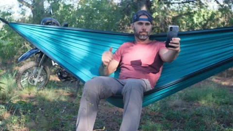 Beard man in a cap is sitting in a hammock in the forest near a motorcycle and d Video stock 214313677