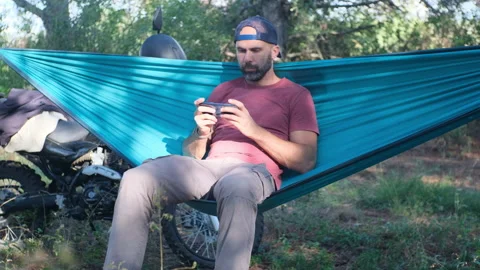 Beard man in a cap is sitting in a hammock in the forest near a motorcycle and w Video stock 214314259