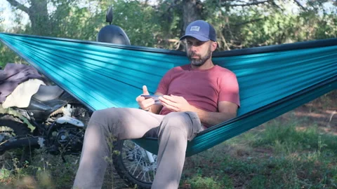 Beard man in a cap is sitting in a hammock in the forest near a motorcycle and w Video stock 214315390