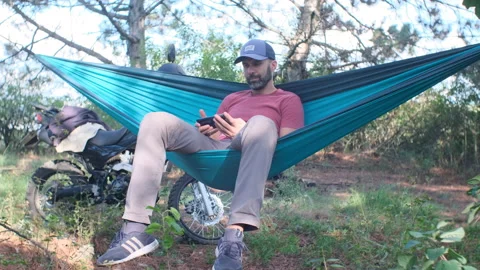 Beard man in a cap is sitting in a hammock in the forest near a motorcycle and w Video stock 214315509