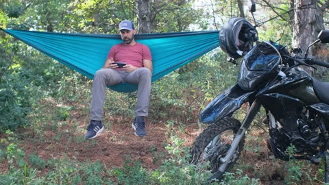 Beard man in a cap is sitting in a hammock in the forest near a motorcycle and w Video stock 214317638