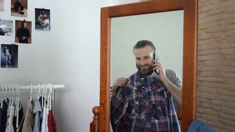 Beard man is looking at himself into a mirror closing a shirt Talking on phone Stock Footage 147894042
