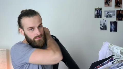 Beard man in a T-shirt  is looking in a mirror putting on his Leather Jacket Stock Footage 147897692