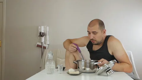 A bearded alcoholic sits at home at a table with a glass of vodka and a bowl, a Stock Footage 142028003