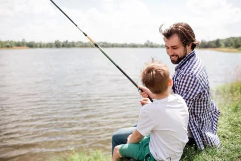 Bearded and handsome guy is sitting with his son at the edge of river shore and Stock Photos