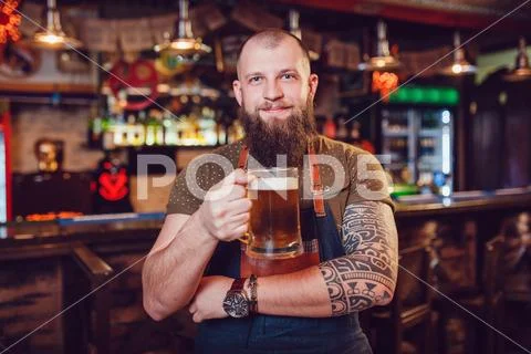 Bearded barman with tattoos wearing an apron standing near the bar and holding a Stock Photos