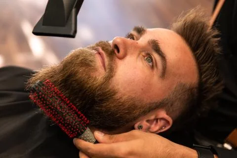 A bearded boy has his beard combed by a barber with a comb and a hairdryer. Stock Photos