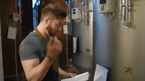 Bearded brewer man on a background of beer tanks and a brewery making notes in a Stock-Footage 99976313