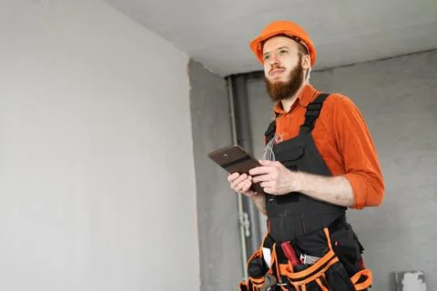 Bearded builder in hardhat and overalls using digital tablet for home renov.. Stock Photos