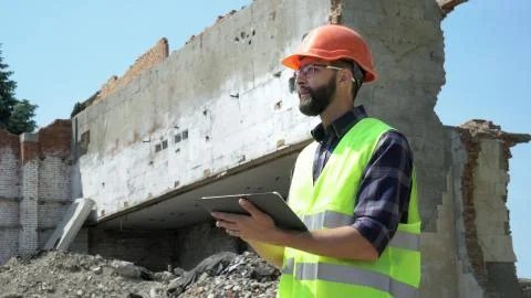 Bearded builder in a helmet and reflective vest uses a tablet on the background Stock Photos