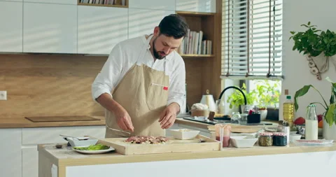 Bearded chef in apron dressing homemade pizza in modern light kitchen with large Stock Footage 218640178