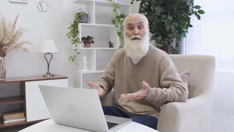 Bearded elderly man talks to camera sitting in soft armchair near laptop Stock Footage 227179348