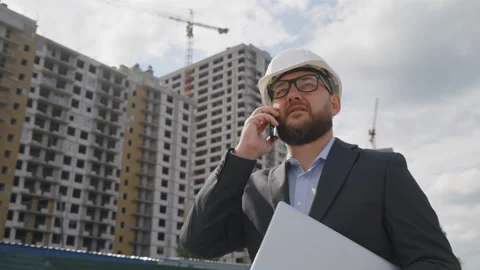 Bearded engineer walking with laptop and talking on phone at construction site Stock Footage 219459663