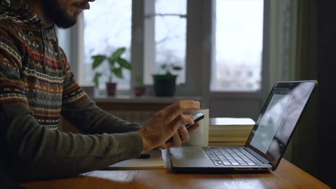 Bearded freelancer is looking in smartphone in front of laptop at his home while Stock Footage 127257236
