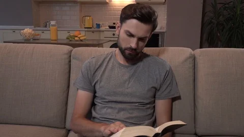 Bearded guy sits on sofa and reads thick book. He turns on pages. Then man looks Stock Footage 99739334