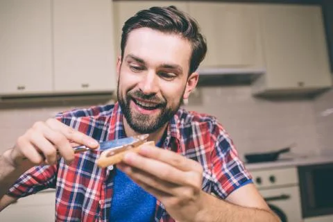 Bearded guy sits at table and holds toast with knife. He puts some butter on it Foto stock